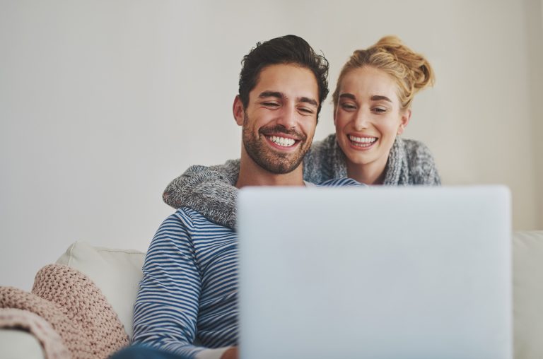 Happy couple sitting on a couch, smiling while looking at a laptop screen, representing financial planning and managing personal loans online.